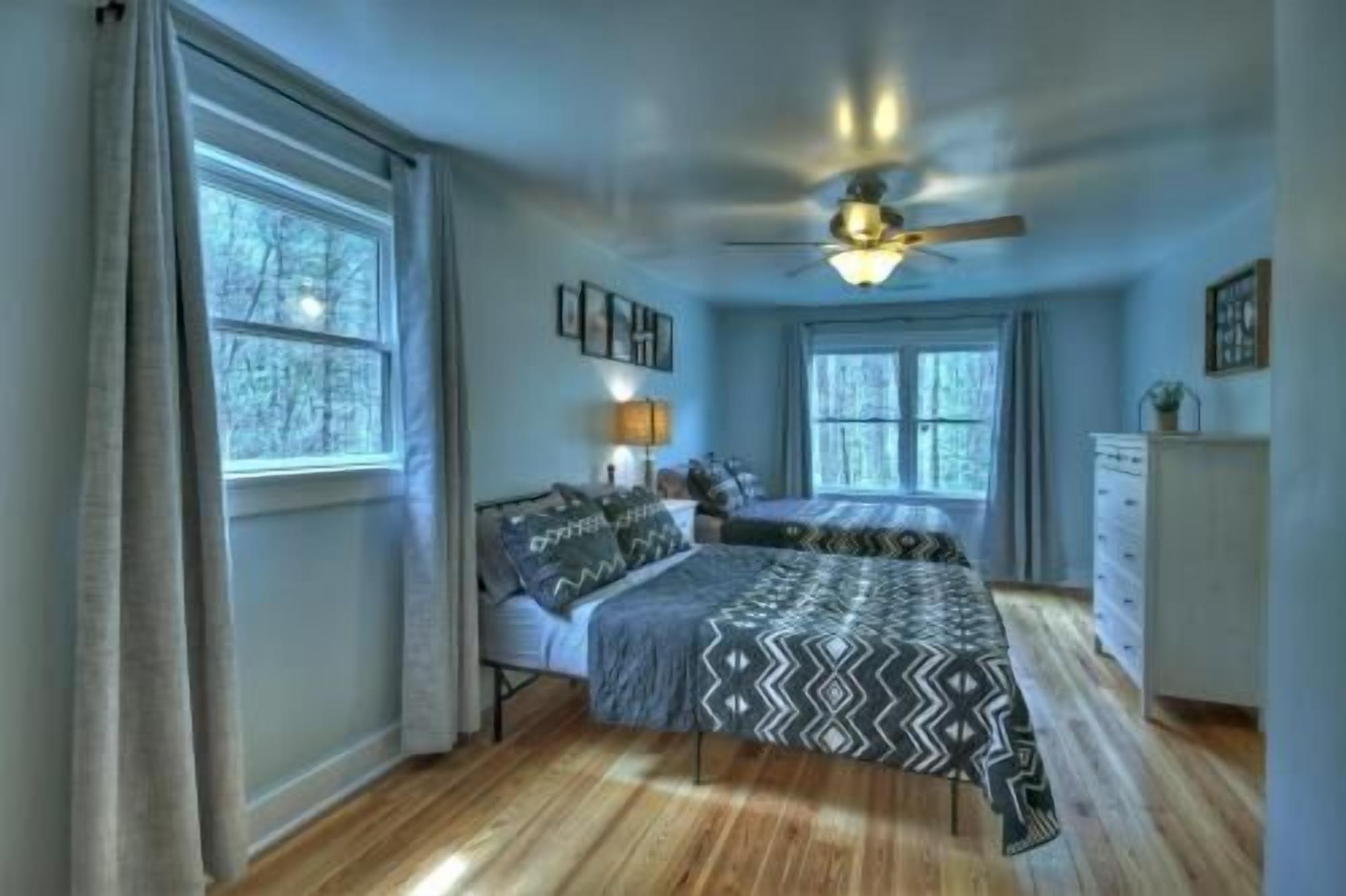 A bedroom with a bed, a ceiling fan, a dresser, and large windows.