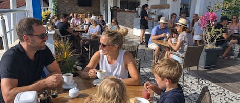 An outdoor café with tables and chairs, a counter with a menu, and a clock on the wall.
