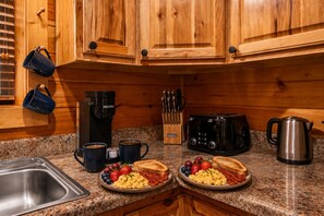 A kitchen with wooden cabinets, a coffee maker, a toaster, a kettle, and breakfast plates with toast, eggs, and fruit. - Cozy Cabin near Pigeon Forge and Gatlinburg, Hot Tub, Fire Pit & Arcade (Sevierville)