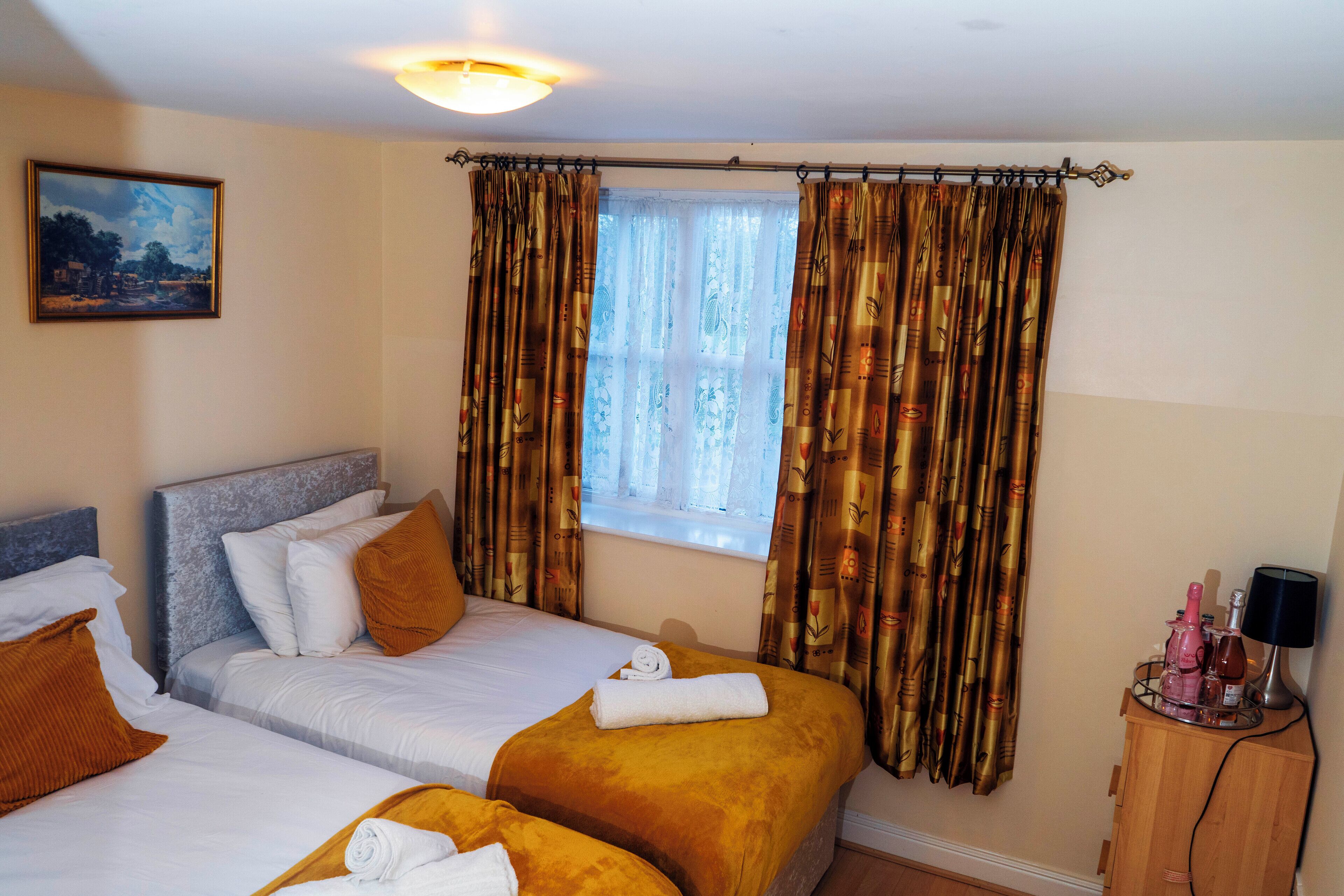 A hotel room with two beds, a window with curtains, a painting on the wall, and a bedside table with a lamp and bottles.