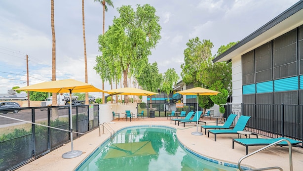A swimming pool with lounge chairs and umbrellas, surrounded by palm trees and a building with blue accents. - Dwell 1723-1 (Scottsdale)