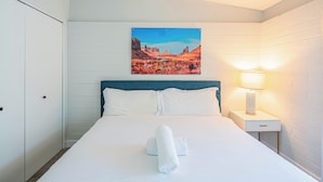A neatly made bed with a rolled towel, a painting of a desert landscape, a bedside lamp, and a white nightstand. - Dwell 1723-1 (Scottsdale)