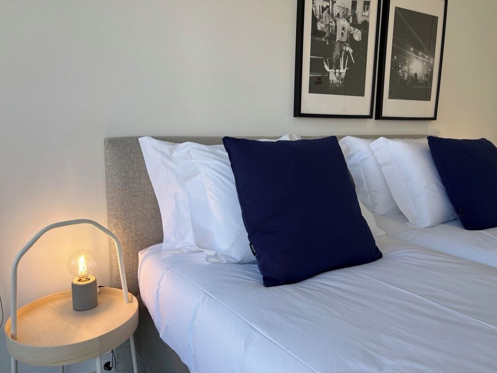 En pænt redt seng med hvide og marineblå puder, et rundt natbord med en lampe, og to billeder i rammer på væggen.