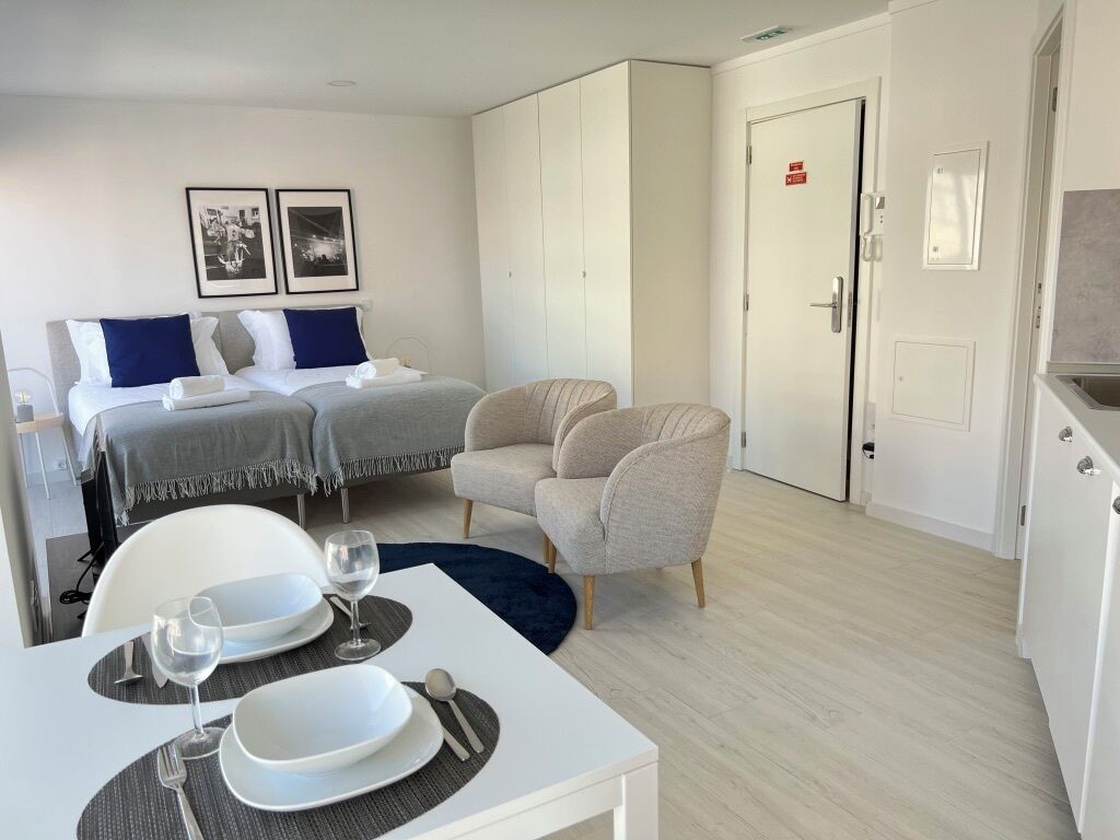 A modern hotel room with a bed, a dining table set with plates and glasses, a chair, and a kitchenette.