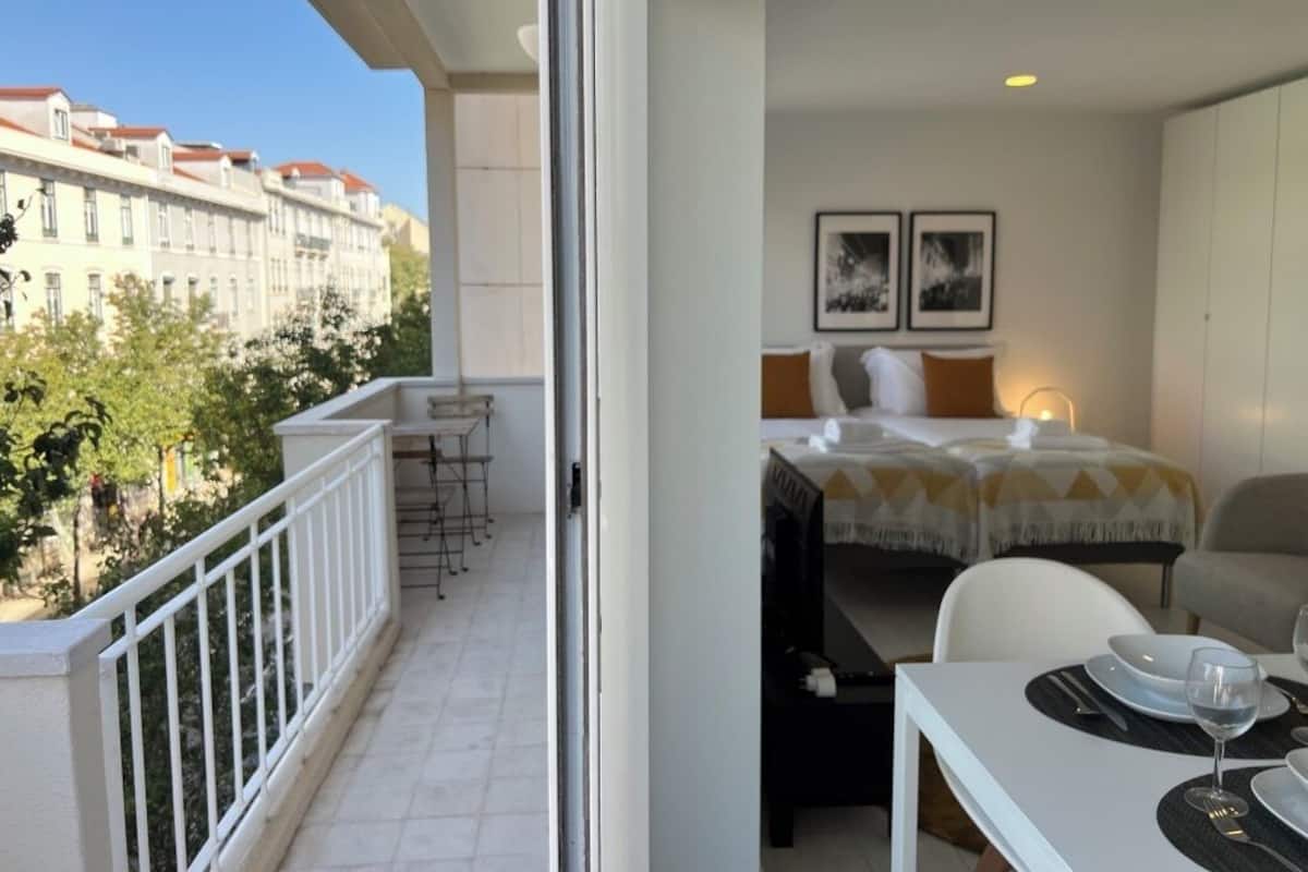 A hotel room with a balcony, a bed with a white and yellow patterned bedspread, a small table with a wine glass, and two framed pictures on the wall.