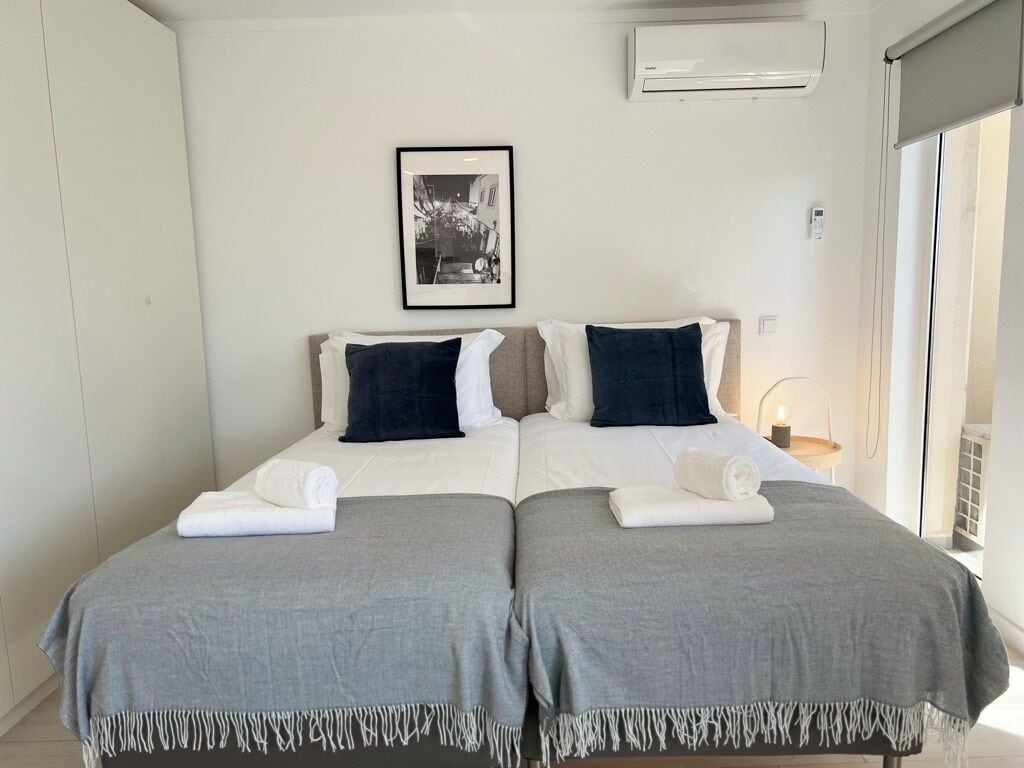 Ein Doppelzimmer mit zwei Betten, jedes mit einer grauen Tagesdecke und weißen Kissen. An der Wand hängt ein gerahmtes Bild, ein Nachttisch mit Lampe und ein Fenster mit Blick.