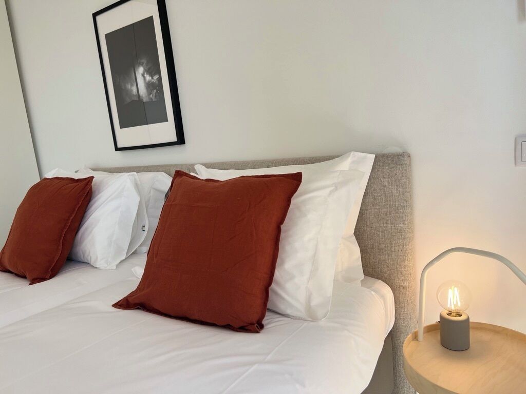 Una cama bien hecha con almohadas blancas y marrones, un cuadro en la pared y una lámpara de noche.
