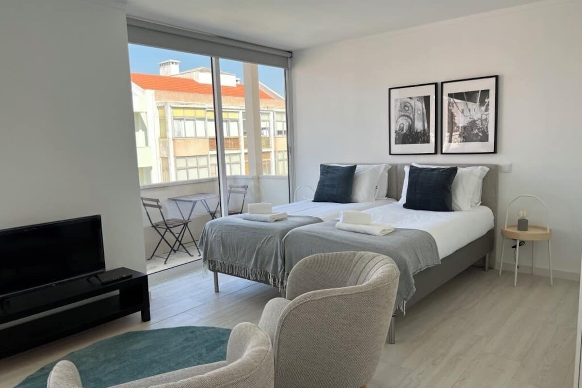A modern bedroom with a bed, two bedside tables, a TV, a chair, and a balcony with a view of buildings.