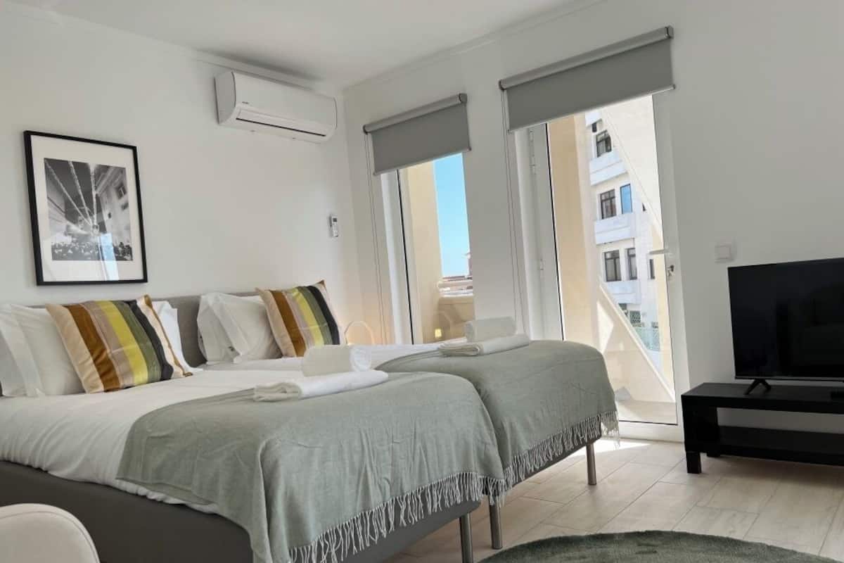 A modern bedroom with a large bed, a TV, and a balcony with a city view.