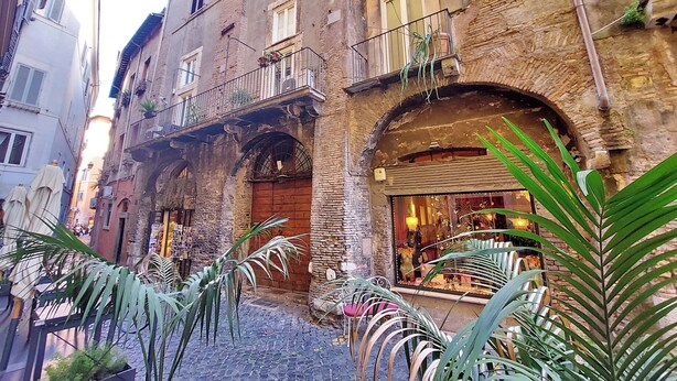A narrow cobblestone street with a building featuring a wooden door, balconies with plants, and a shop with glass display. - Piazza Navona Hydromassage Tub 7 people (Rome)