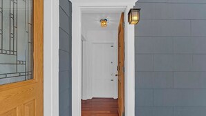 Un pasillo estrecho con una puerta de madera a la izquierda, una puerta blanca al final y una lámpara arriba.