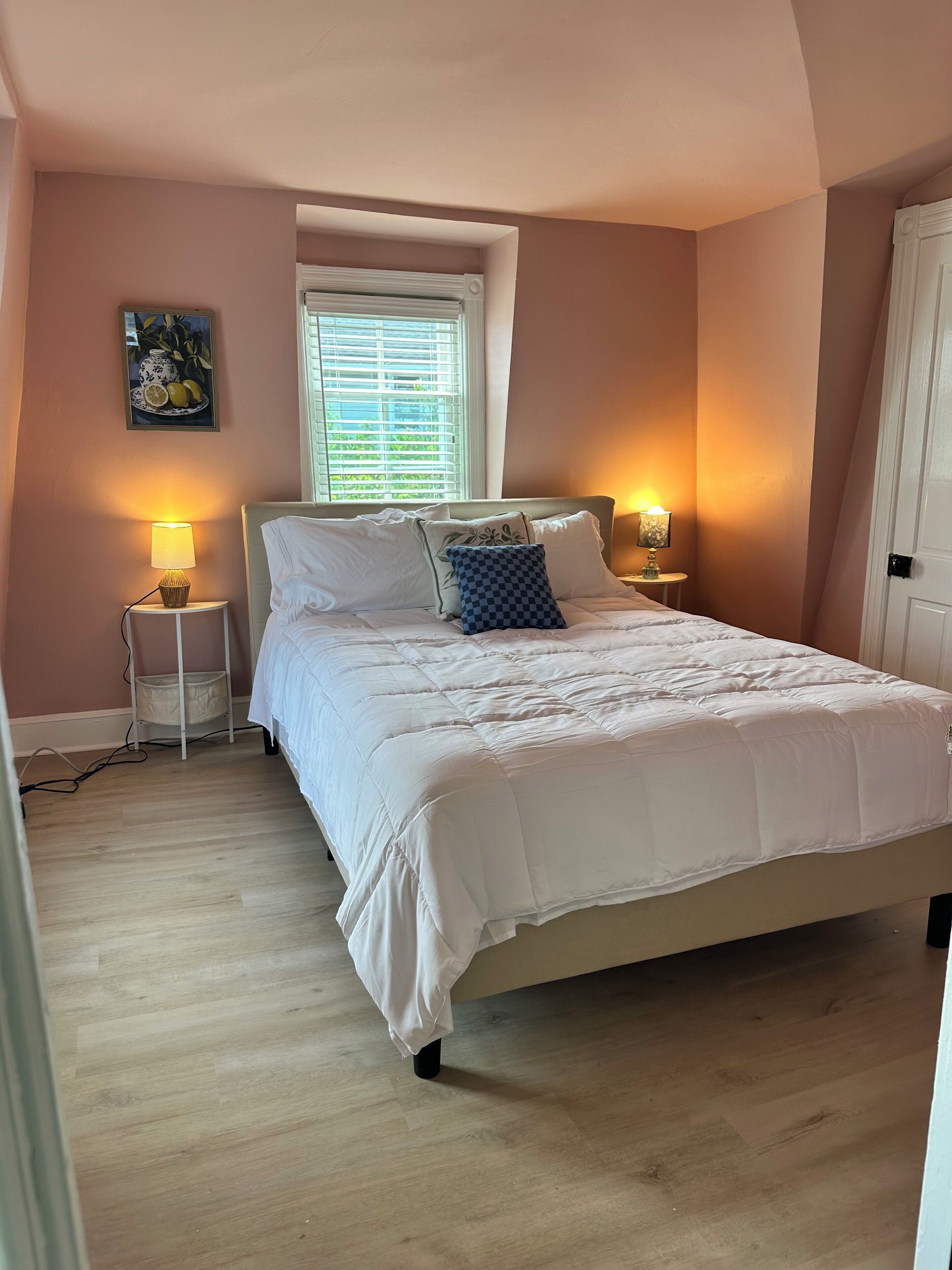Une chambre à coucher comprenant un lit, deux lampes de chevet, une fenêtre avec des stores, un tableau accroché au mur et une porte.