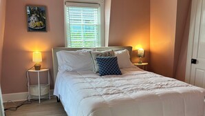 Un dormitorio con una cama, dos lámparas de noche, una ventana con persianas, un cuadro en la pared y una puerta.
