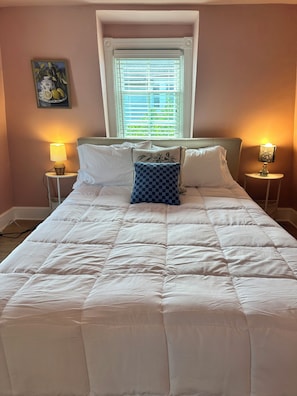 A neatly made bed with a checkered pillow, two bedside lamps, and a window with blinds.