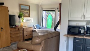 Sala de estar con un sofá de cuero marrón, una mesa de centro de madera y un televisor sobre un mueble de madera. Hay una zona de cocina con armarios blancos y un microondas. Se ve una tabla de surf a través de una ventana.
