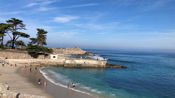 Una playa con un muro de piedra, un muelle y gente caminando por la orilla.