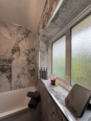 Baño con pared de mármol, bañera, ventana con vidrio satinado y artículos de aseo en el alféizar.
