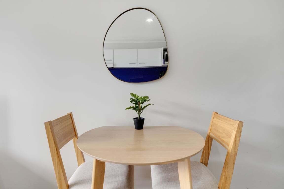 Une table ronde en bois, accompagnée de deux chaises, d’une plante en pot et d’un miroir fixé au mur.