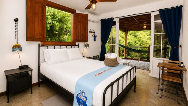 Un dormitorio con cama, mesita de noche, escritorio, hamaca y vistas al exterior con vegetación.