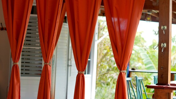 A room with orange curtains tied back, revealing a window with blinds and a view of greenery outside. - Private Finca Ki' •3BR • Hammock Terrace • El Yunque • Ceiba Ferry • Easy-Access (NAGUABO)