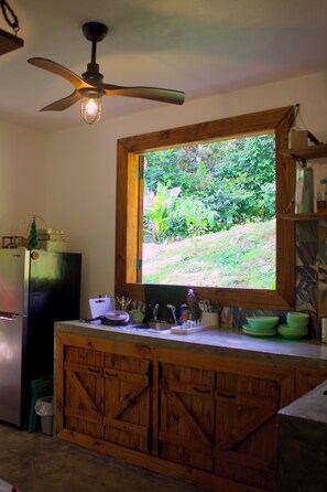 Une cuisine avec des meubles de rangement en bois, un réfrigérateur, une fenêtre donnant sur un espace verdoyant et un ventilateur de plafond.