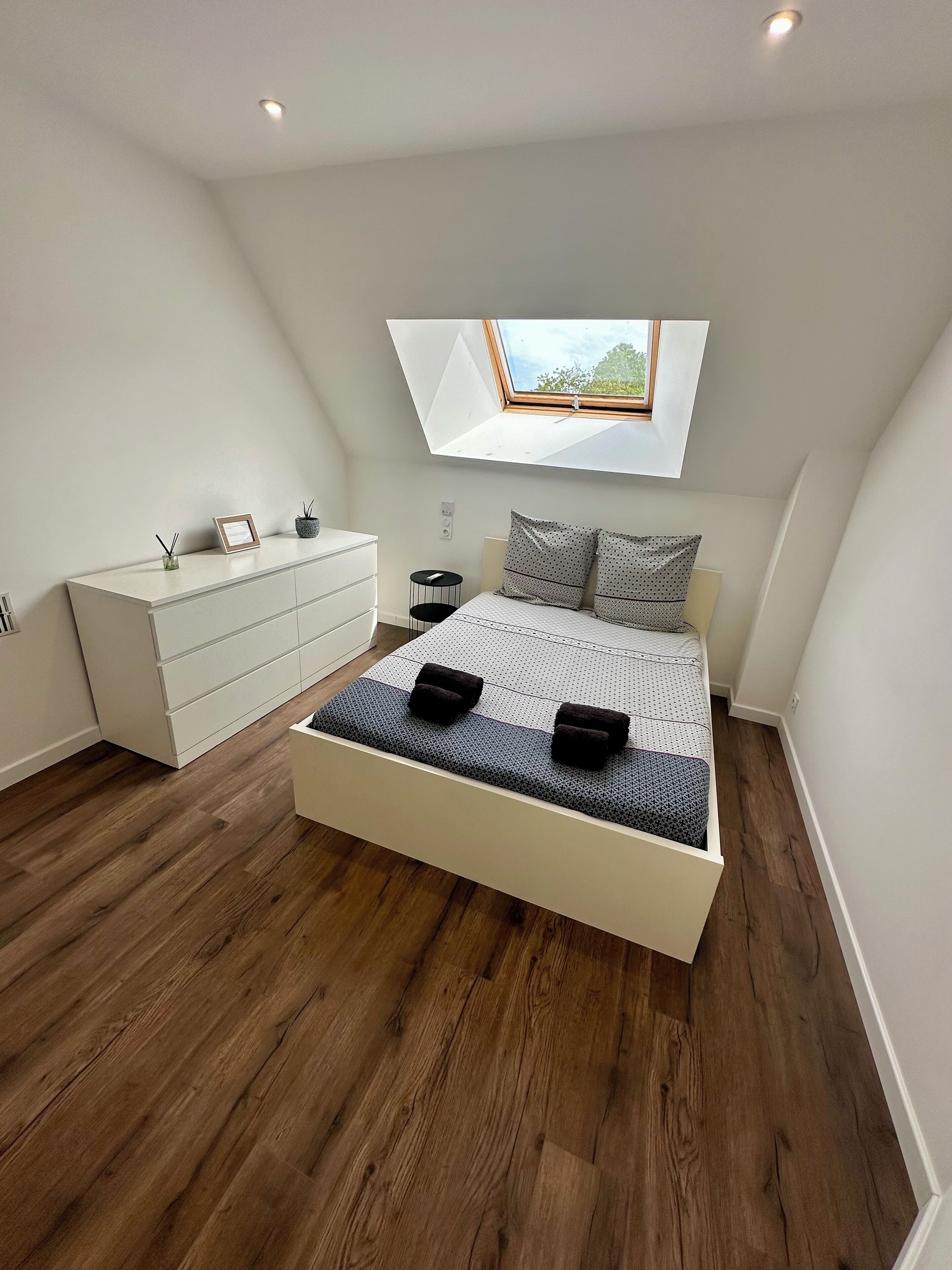 Ein modernes Schlafzimmer mit schrägem Dach, einem Oberlicht, einem großen Bett mit gemustertem Überwurf, einer weißen Kommode und einem kleinen Nachttisch.