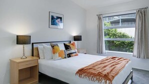Una camera d'albergo moderna con un letto grande, due comodini, un tavolino e una finestra con tende. Si vede anche un quadro appeso al muro.