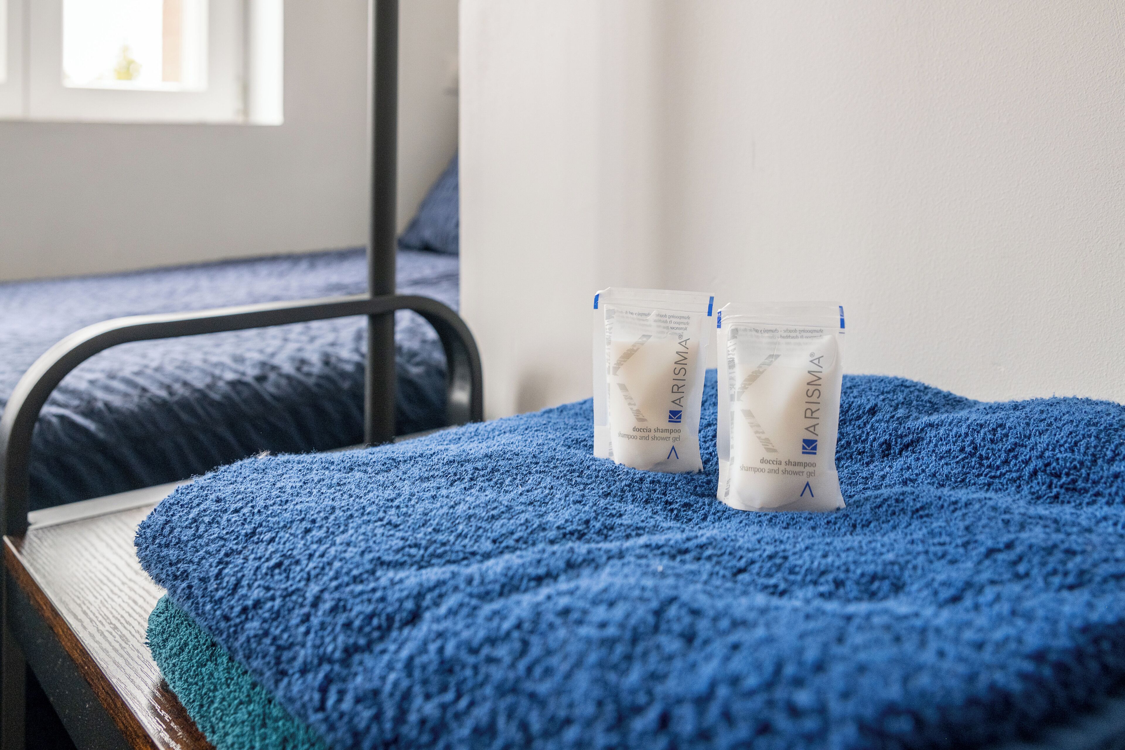 Sängyllä on sininen pyyhe ja kaksi pakettia Arisia-shampoota.