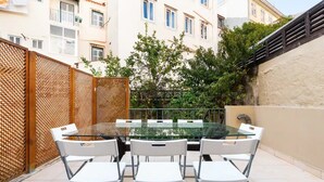 A glass-top table with white chairs in an outdoor patio area, surrounded by a wooden fence and buildings in the background. - Elegant One-Bedroom House• Terrace & Garage (Lisboa)
