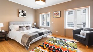 A modern bedroom with a large bed, bedside tables, a colorful rug, and a view of buildings through large windows. - Elegant One-Bedroom House• Terrace & Garage (Lisboa)