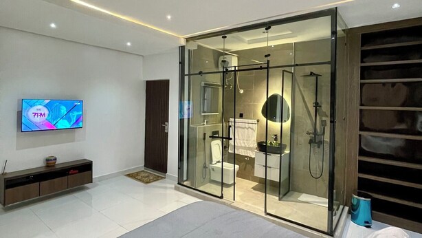 A modern bedroom with a large bed, a TV mounted on the wall, and a glass-enclosed bathroom with a shower and toilet.