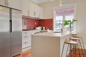 A modern kitchen with a stainless steel refrigerator, white cabinets, a red backsplash, a gas stove, a sink, and bar stools.