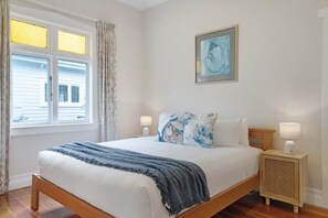 A bedroom with a bed, a nightstand, a lamp, a window with curtains, and a painting on the wall.
