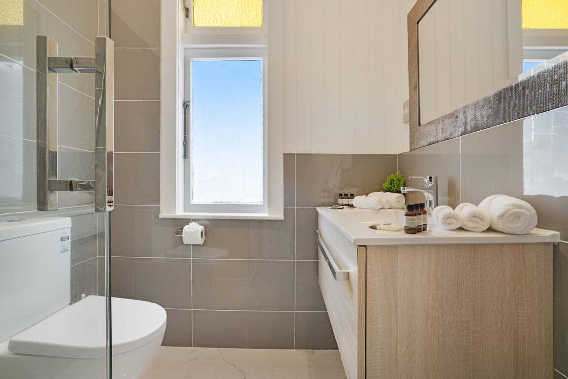 A modern bathroom with a glass shower enclosure, a toilet, a sink with toiletries, and a window providing natural light.