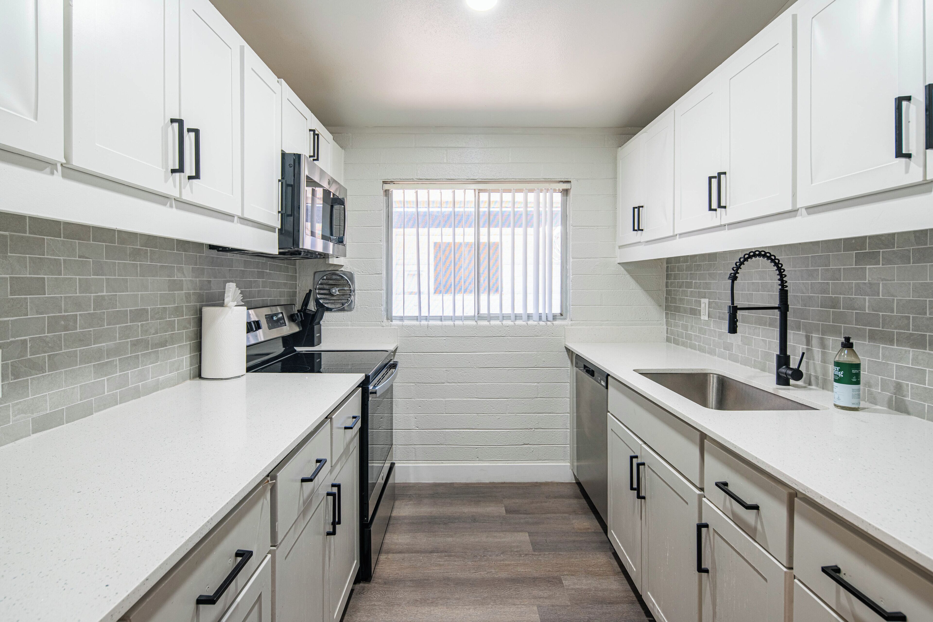 A modern kitchen with white cabinets, grey tiled backsplash, stainless steel appliances, and a window with blinds.