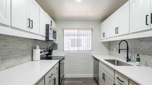 Een moderne keuken met witte kasten, een grijze betegelde spatwand, roestvrijstalen apparatuur en een raam met jaloezieën.