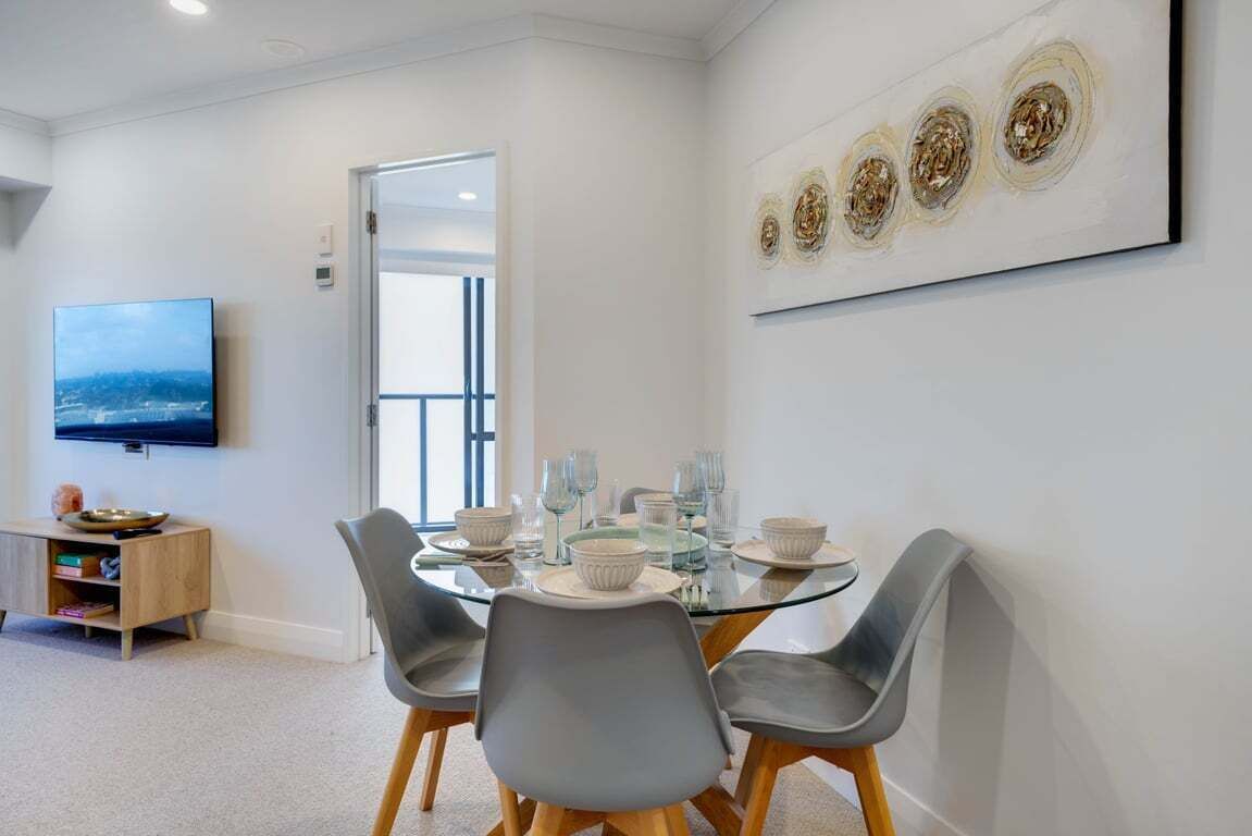 A dining area with a glass table, grey chairs, and a television on the wall.