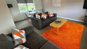 A modern living room with a grey sofa, orange and white patterned cushions, a wooden coffee table, and a vibrant orange rug. - Apartment - Sleeps4 - Glasgowairport 10 Mins Drive (Renfrew)
