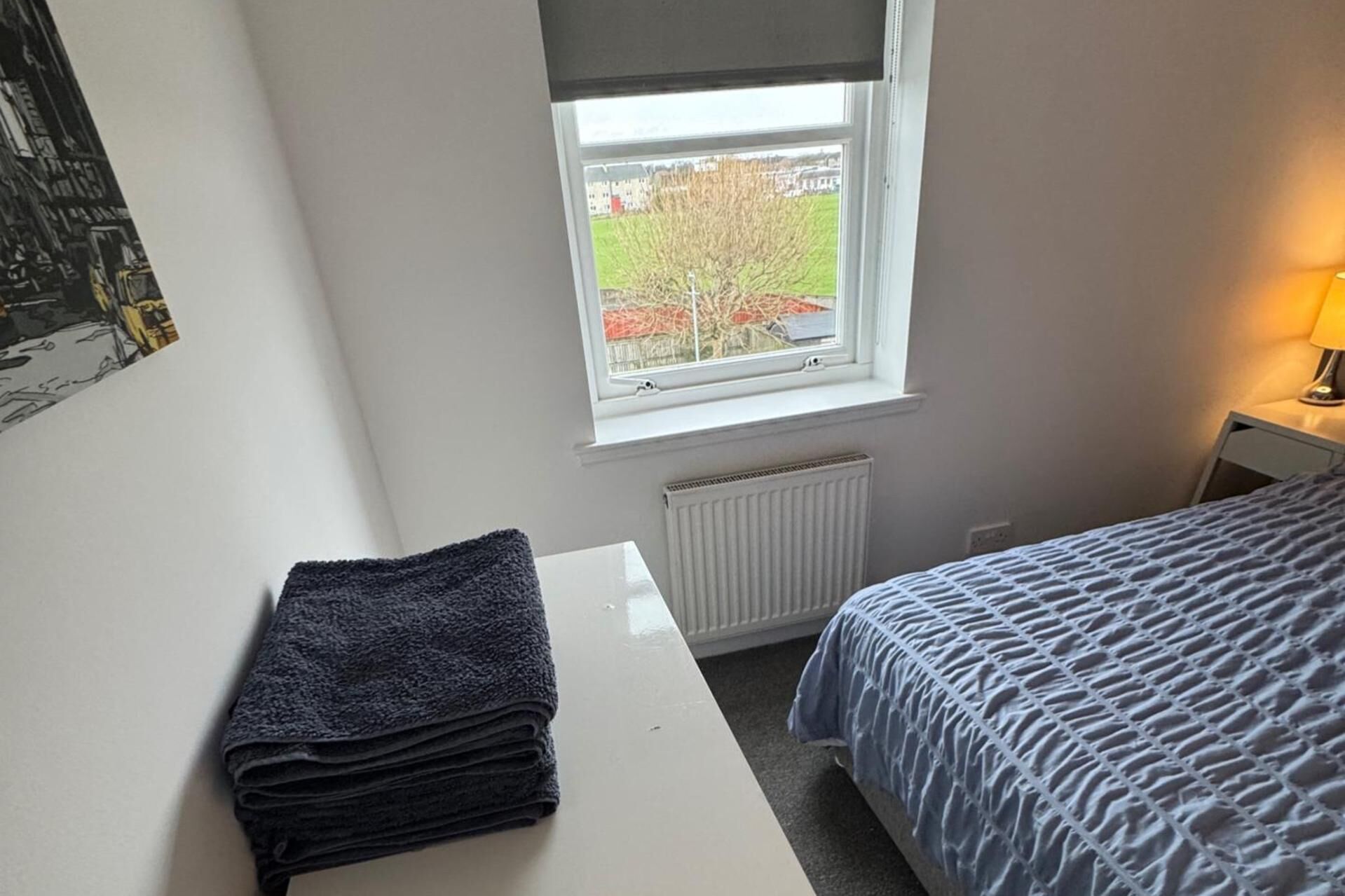 A bedroom with a bed, a radiator, a window with a view of trees and buildings, and a stack of towels on a table.