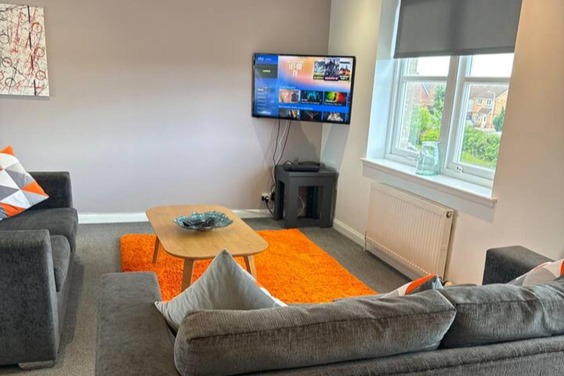 Uma sala de estar moderna com um sofá cinzento, tapete laranja e uma TV de tela plana fixada na parede.