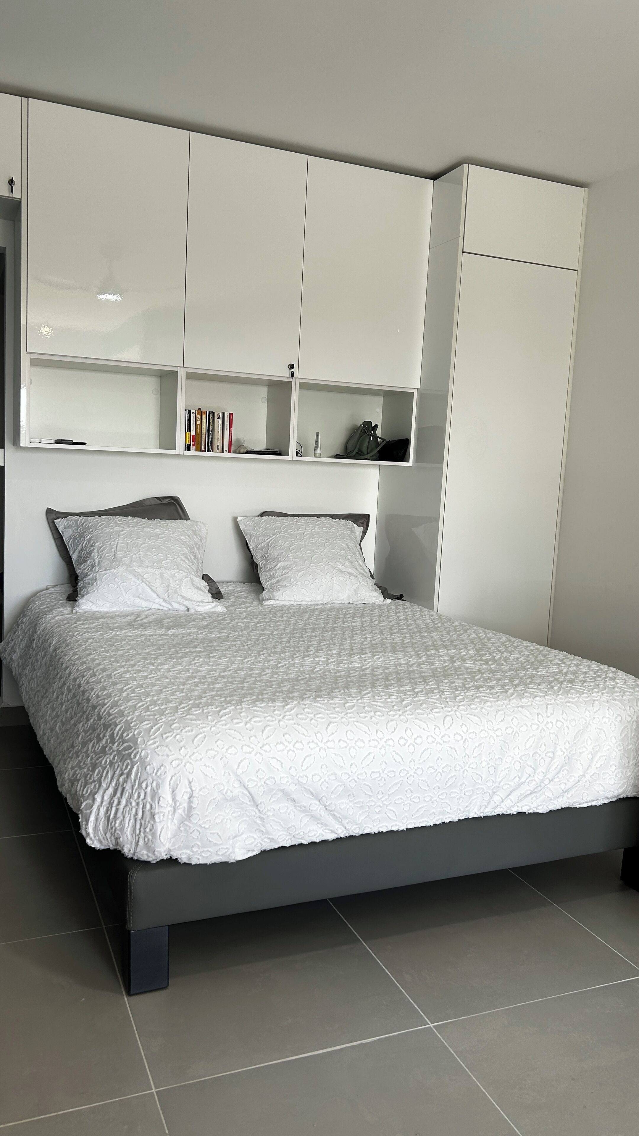 Ein Bett mit weißer Bettdecke und Kissen, ein grauer Kopfteil und weiße, integrierte Regale darüber.