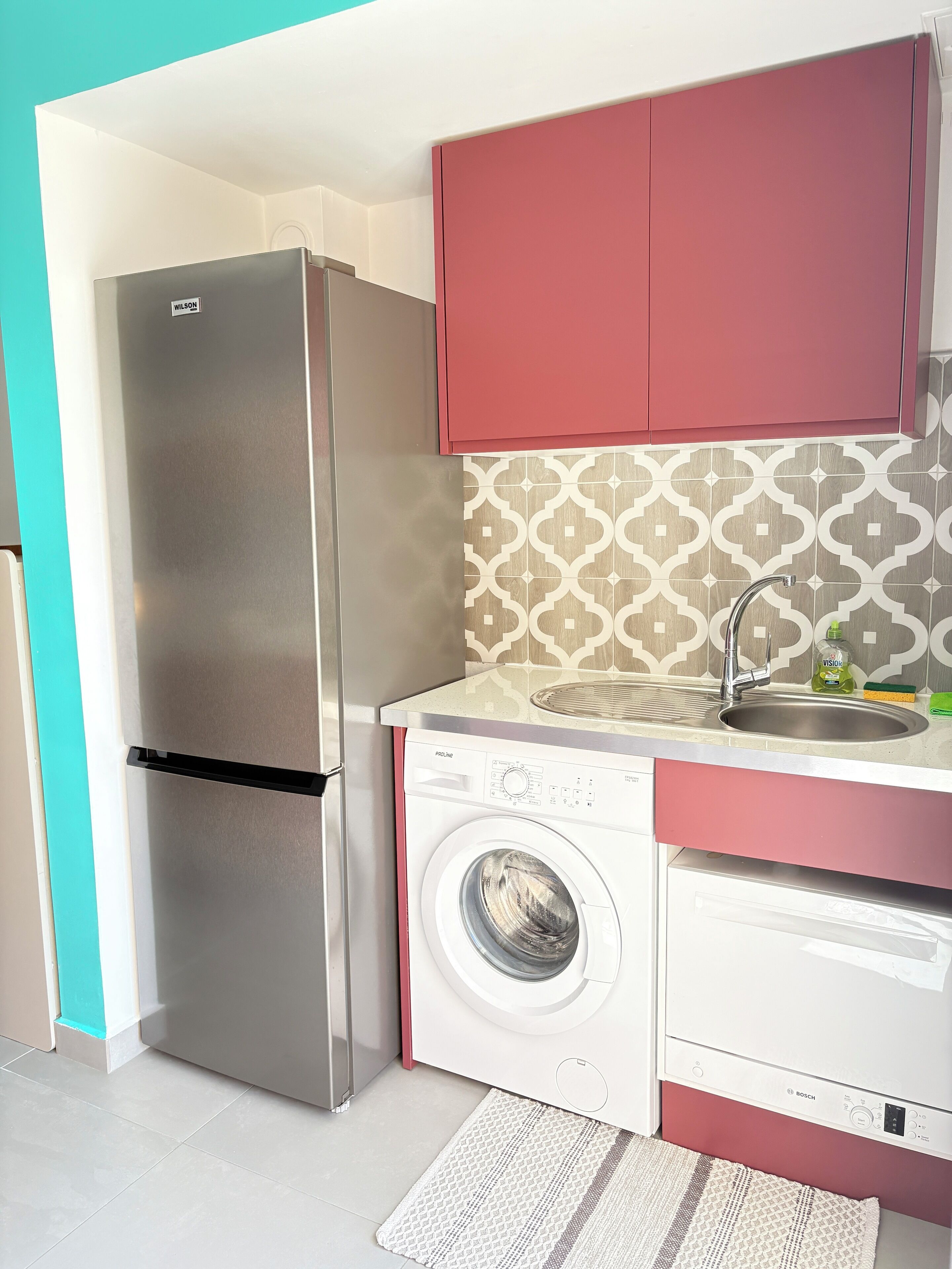A compact kitchen with a stainless steel refrigerator, a Bosch washing machine, and a sink with a patterned backsplash.
