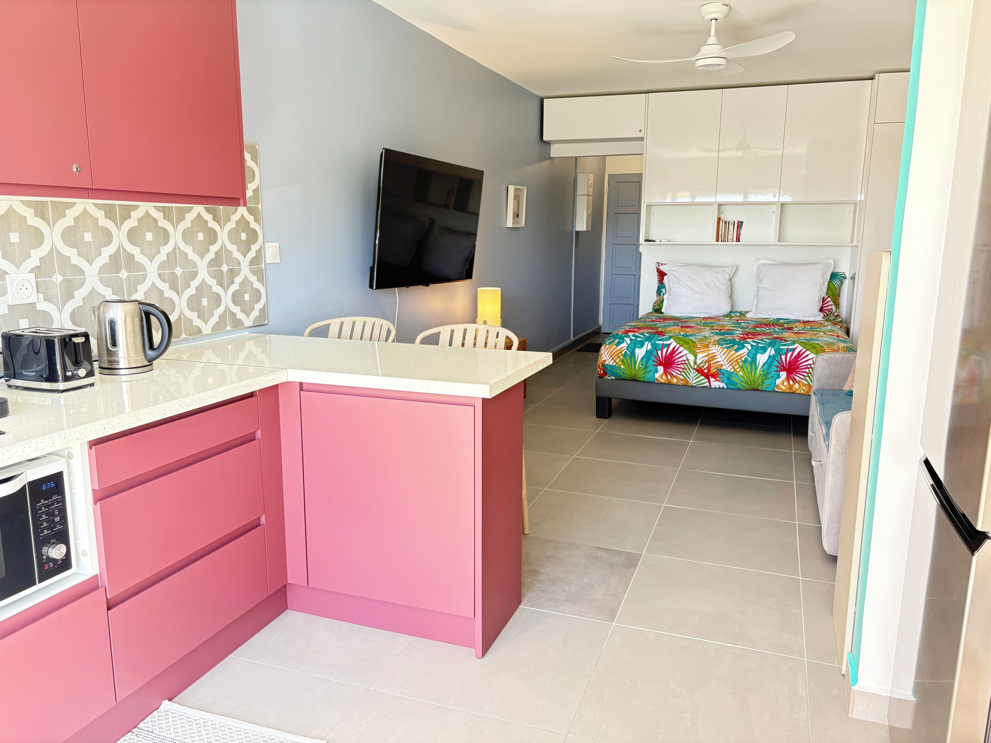 Une chambre d’hôtel compacte dotée d’une kitchenette, d’un lit recouvert d’une courtepointe à motifs floraux colorés, d’un téléviseur à écran plat fixé au mur et d’un ventilateur de plafond.