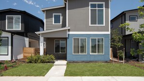 Une maison moderne à deux étages, avec une façade grise et bleue, un jardin devant la maison avec un chemin et un petit arbre.