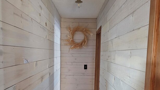 En smal hall med träväggar och ett trägolv, som leder fram till en dörr med en krans på väggen.