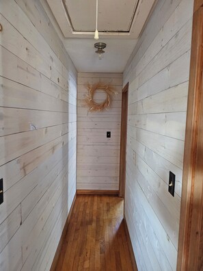 A narrow hallway with wooden walls and a wooden floor, leading to a door with a wreath on the wall. - Perfect 2-bedroom house for EAA 2026 in Omro's quiet setting. (Omro)