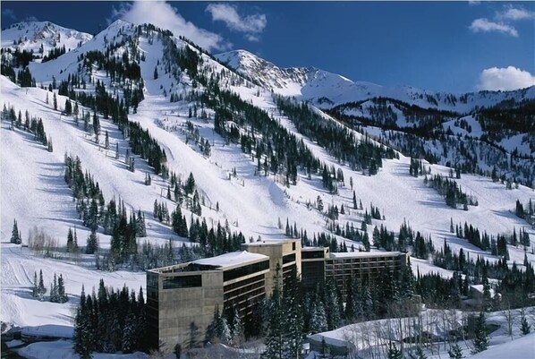 A snow-covered mountain landscape with modern buildings featuring a grid-like facade and large windows, surrounded by dense evergreen trees.
