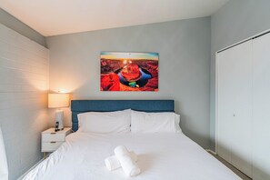 A neatly made bed with white linens and a rolled towel, a bedside table with a lamp, and a wall-mounted painting of a canyon. - Dwell 1911-3 (Scottsdale)