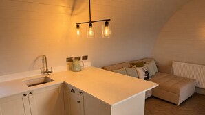 Une cuisine de taille réduite, équipée d’un évier, de placards et d’un plan de travail. On aperçoit un canapé avec des coussins et un radiateur à l’arrière-plan.