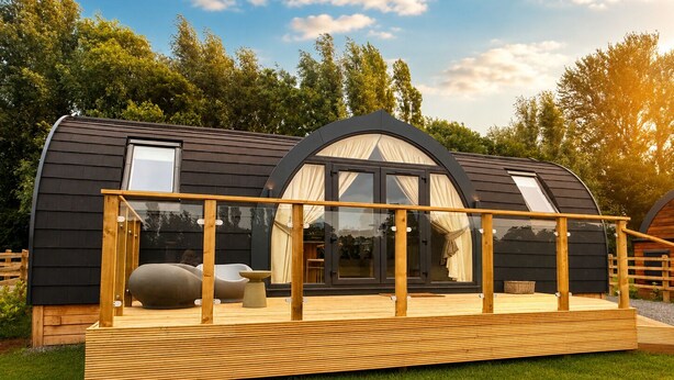 Une cabane moderne en bois aux lignes courbes, dotée d’une terrasse, de grandes fenêtres et d’une rambarde en verre.
